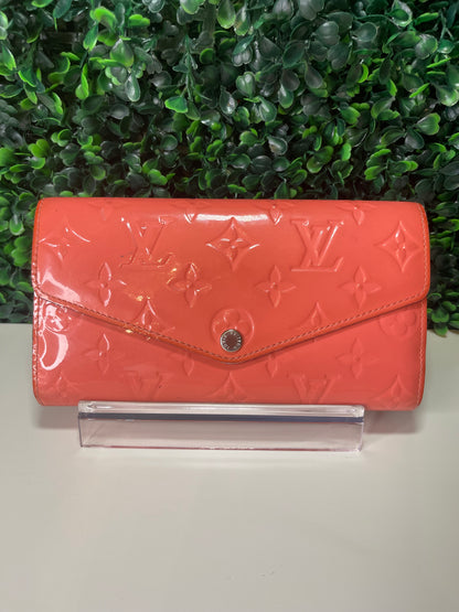 Front view of a pink glossy preowned Louis Vuitton long wallet with vernis leather and a front flap snap, silver hardware button snap, displayed on a clear stand.