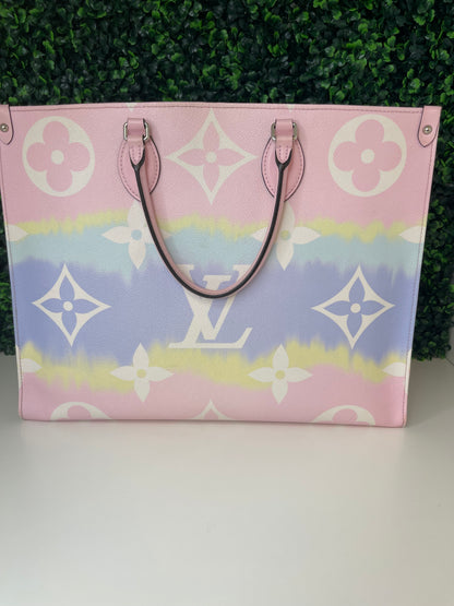A pink and multicolor patterned Louis Vuitton OnTheGo Escale handbag GM with a structured handle and straps, displayed against a green leafy backdrop.