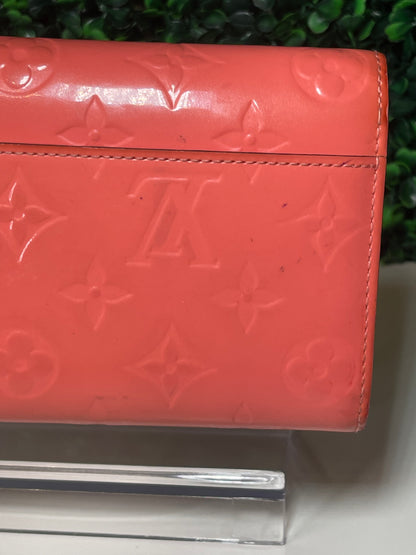 Back side view of a pink glossy preowned Louis Vuitton long wallet with vernis leather and a front flap snap, silver hardware button snap, displayed on a clear stand. Some light ink marks observed on back side. 