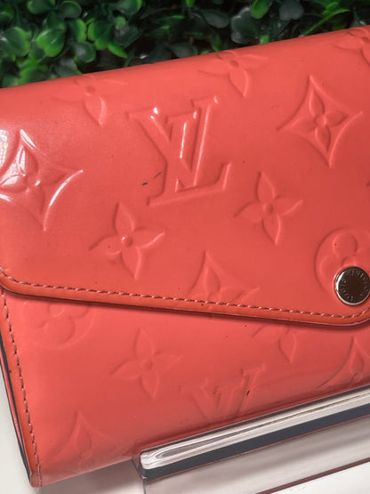 Front view of a pink glossy preowned Louis Vuitton long wallet with vernis leather and a front flap snap, silver hardware button snap, displayed on a clear stand. Shows small ink mark on front of wallet. 