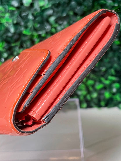 Side view of a pink glossy preowned Louis Vuitton long wallet with vernis leather and a front flap snap, silver hardware button snap, displayed on a clear stand.