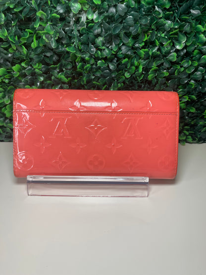 Back view of a pink glossy preowned Louis Vuitton long wallet with vernis leather and a front flap snap, silver hardware button snap, displayed on a clear stand.