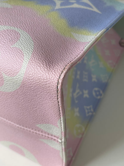 Zoomed in image of bottom corner view of a pink base and multicolor patterned Louis Vuitton OnTheGo Escale handbag GM laying on its side.