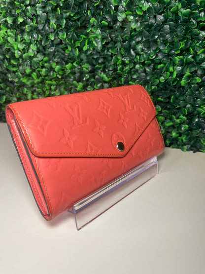 Side view of a pink glossy preowned Louis Vuitton long wallet with vernis leather and a front flap snap, silver hardware button snap, displayed on a clear stand.