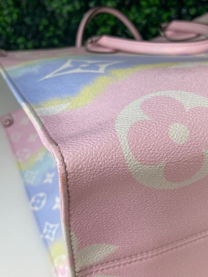 Zoomed in image of bottom corner view of a pink base and multicolor patterned Louis Vuitton OnTheGo Escale handbag GM laying on its side.