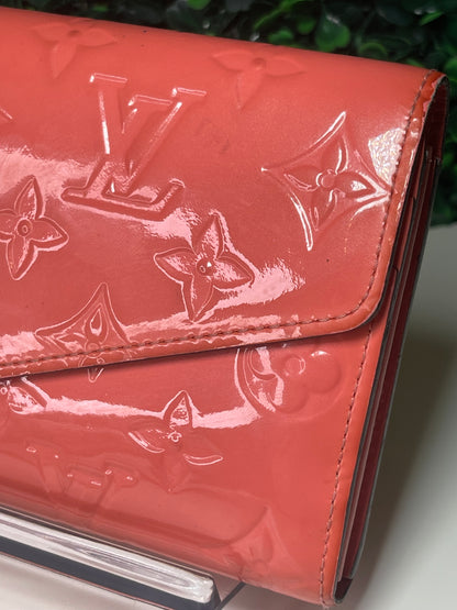 Front view of a pink glossy preowned Louis Vuitton long wallet with vernis leather and a front flap snap, silver hardware button snap, displayed on a clear stand.