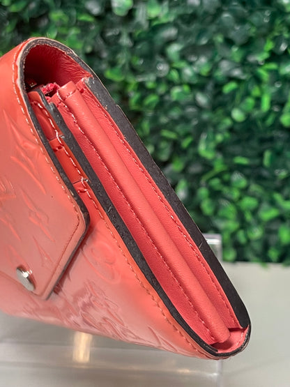 Side view of a pink glossy preowned Louis Vuitton long wallet with vernis leather and a front flap snap, silver hardware button snap. 