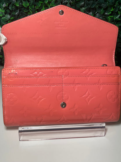 Front view of a pink glossy preowned Louis Vuitton long wallet with vernis leather and a front flap snap opened, card slots in interior, and silver hardware button snap, displayed on a clear stand.